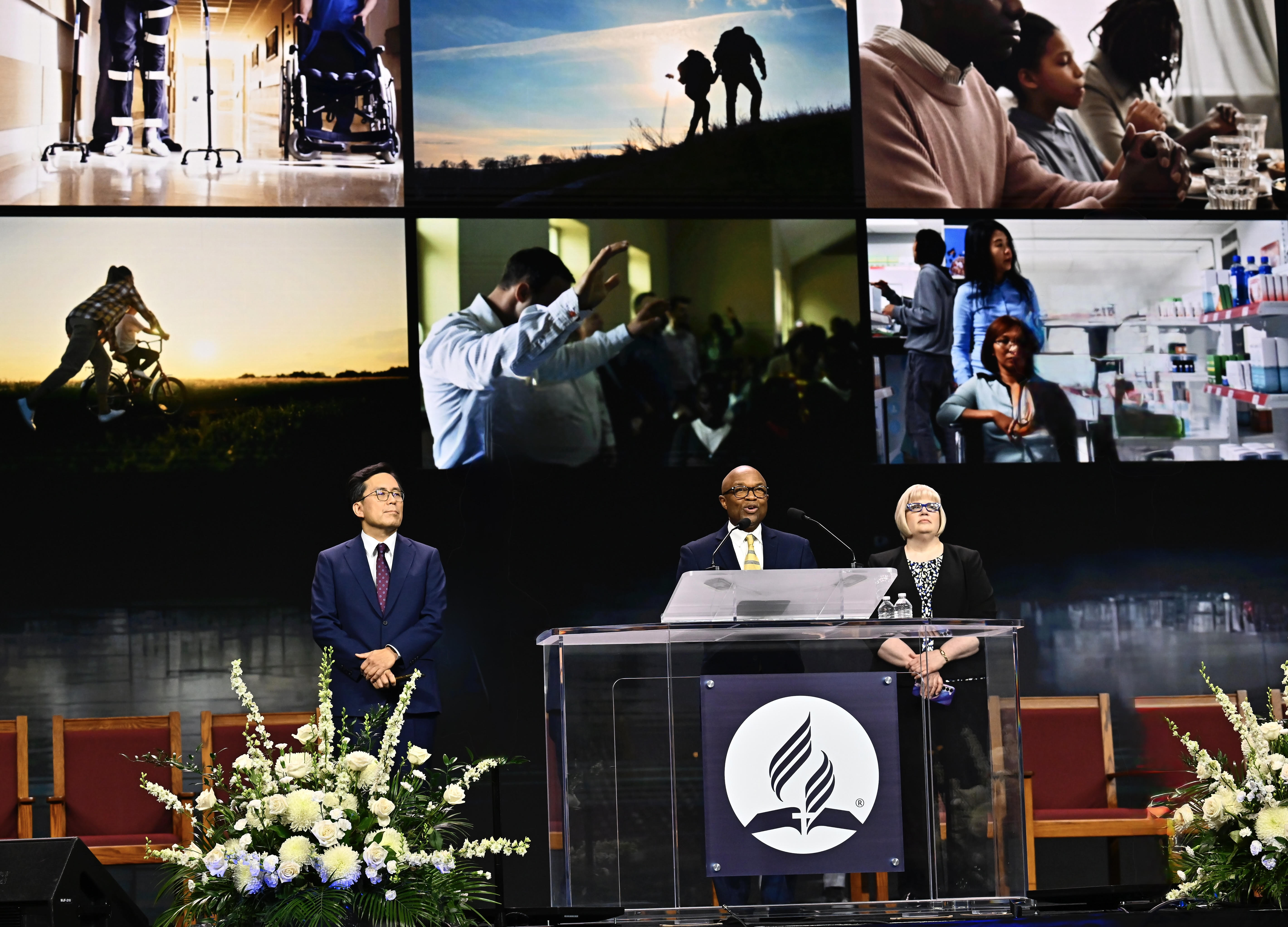 Left to right - an Asian man, black man, and white woman stand on a stage with a collage of ministry pictures behind them. The black man speaks at the podium.